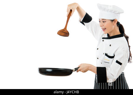 Bella chef titolari di una spatola di legno in aria che mostra il gesto di cottura con copyspace e tenendo un pan sorriso in piedi su un muro bianco sullo sfondo. Foto Stock