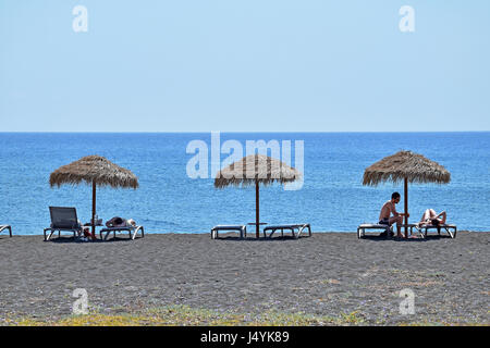 Tourist in appoggio sui lettini sotto gli ombrelloni di paglia a Perissa, isola di Santorini, Cicladi Grecia Foto Stock