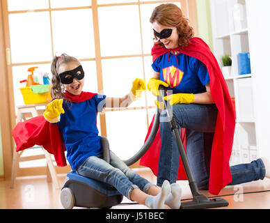 Il bambino e la madre vestita come supereroi utilizzando aspirapolvere in camera. Famiglia di mezza età della donna e la figlia hanno un divertimento durante la pulizia del pavimento. Foto Stock