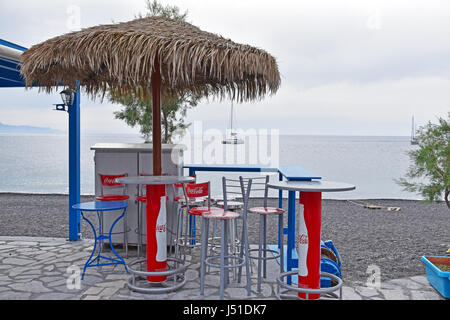 Ombrelloni di paglia su tavoli e sedie pubblicità coca cola su Kamari beach - Santorini, Grecia Foto Stock