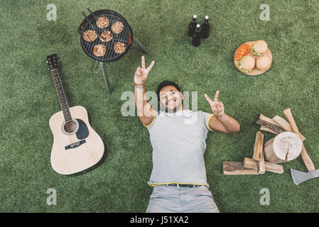 Vista dall'alto di felice giovane uomo appoggiato su erba con la chitarra e cibo per picnic Foto Stock