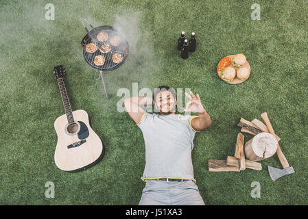 Vista dall'alto di felice giovane uomo che mostra segno ok giacente su erba con la chitarra e cibo per picnic Foto Stock