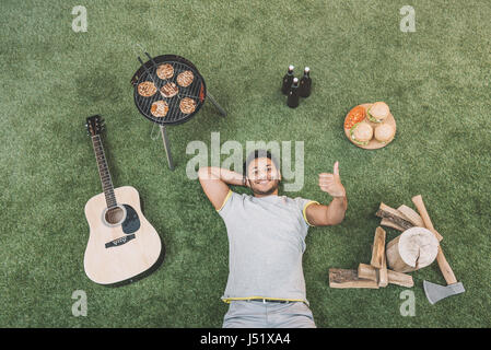 Vista dall'alto di felice giovane uomo che mostra un pollice alzato e poggiante su erba con la chitarra e cibo per picnic Foto Stock