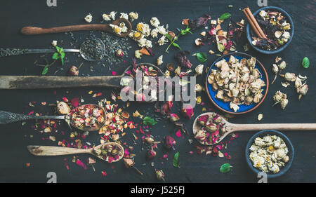 Cucchiai di legno secco con erbe aromatiche, boccioli di fiori e foglie di tè sul nero bruciata sullo sfondo di legno, vista dall'alto, composizione orizzontale Foto Stock