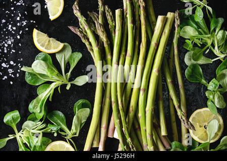 Bundle di giovani di materie organiche non cotte asparagi verdi con insalata verde foglie, fette di limone e il Sale marino nero su sfondo di texture. Vista dall'alto. Healt Foto Stock