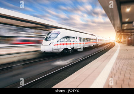 Bianco moderno treno ad alta velocità in movimento sulla stazione ferroviaria al tramonto. Treno sul binario ferroviario con motion blur effetto in Europa in serata. Stazione ferroviaria pl Foto Stock