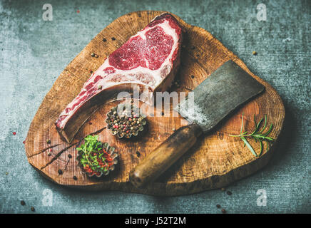Invecchiato a secco di manzo crudo costata di manzo con osso, butcher carne lama per tritare e spezie in ciotole rustico di legno su cemento grigio Sfondo, top v Foto Stock