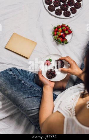 Una donna con i jeans sedersi e mangiare i biscotti e fragole fresche. Un vecchio libro, una piastra di fragole fresche e accompagnare i cookie. Foto Stock