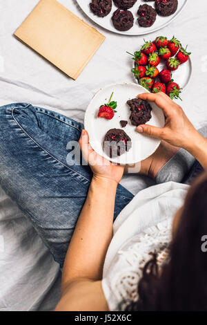 Una donna con i jeans sedersi e mangiare i biscotti e fragole fresche. Un vecchio libro, una piastra di fragole fresche e accompagnare i cookie. Foto Stock