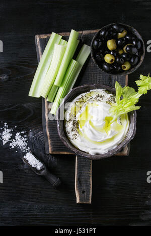 Sedano fresco con yogurt e olio d'oliva tuffo nella ciotola in ceramica, servita con sale marino e nero, olive verdi su legno tagliere su legno nero masterizzare Foto Stock