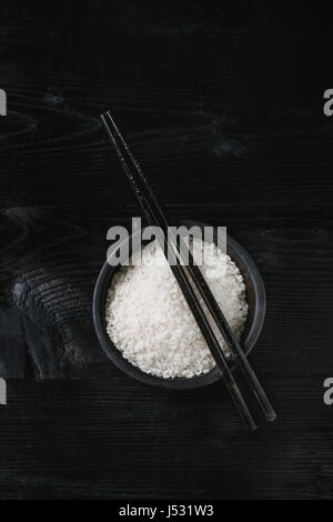 Bianco crudo sushi il riso crudo in nero ciotola con bacchette oltre bruciati sullo sfondo di legno. Vista da sopra con lo spazio di copia Foto Stock