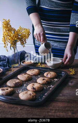 Donna versando latte di mandorla in una tazza e nocciola biscotti con scaglie di cioccolato su un vassoio da forno Foto Stock