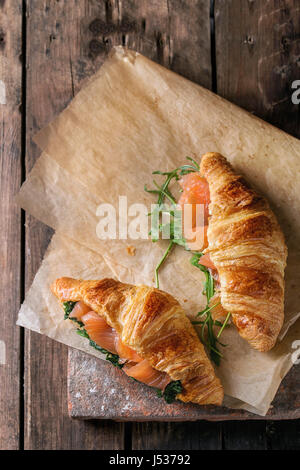 Due croissant con affumicati Salmoni salati, spinaci e rucola servita sulla carta da forno di terracotta oltre il bordo scuro legno vecchio sfondo. Vista da sopra con sp Foto Stock