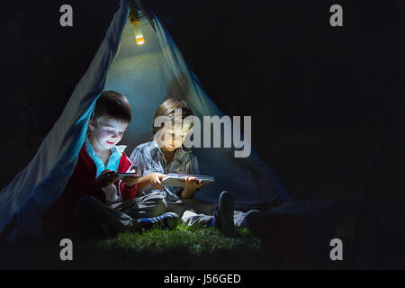 Carino piccoli fratelli, giocando su tablet e telefono di notte in campside, nella tenda Foto Stock