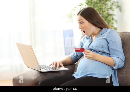 Stupiti donna incinta acquisti on line con carta di credito e un laptop seduto su un divano nel soggiorno in un interno di una casa Foto Stock