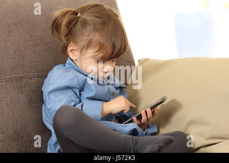 Concentrato baby giochi con uno smart phone on line seduta su un divano nel soggiorno di casa Foto Stock