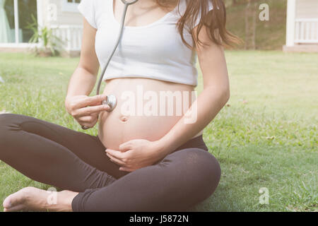 Asian donna incinta ascoltando la sua pancia utilizzando uno stetoscopio sul verde grasss davanti a casa sua. Foto Stock