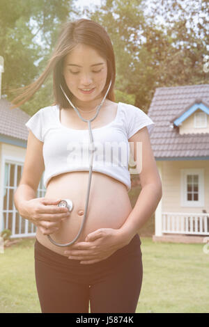 Asian donna incinta ascoltando la sua pancia utilizzando uno stetoscopio sul verde grasss davanti a casa sua. Foto Stock