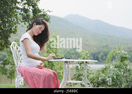 Giovani asiatici donna incinta piani il budget familiare con la calcolatrice. bellissima femmina calcolando la sua bollette domestiche in giardino Foto Stock