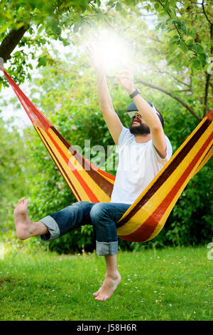 Giovane uomo barbuto che indossa la realtà virtuale occhiali rilassante in un giardino amaca. lifestyle vr divertimento e relax concept Foto Stock