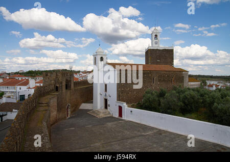 Alta Vista di Alandroal chiesa lungo il castello di mura difensive, il villaggio circostante e la torre dell orologio sotto un azzurro cielo velato. Alentejo, Portogallo Foto Stock