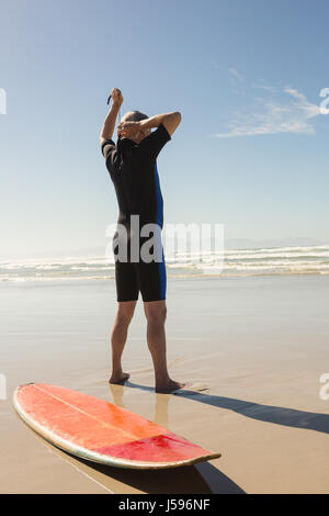 Vista posteriore di uomo che indossa mentre muta in piedi da tavola da surf in spiaggia Foto Stock