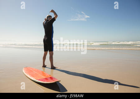 Vista posteriore del senior uomo muta da indossare mentre in piedi da tavola da surf in spiaggia Foto Stock
