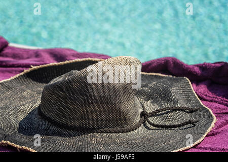 Concetto di estate. La paglia cappello per il sole su un asciugamano viola con un blu sullo sfondo di acqua Foto Stock