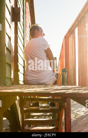 Vista posteriore dell'uomo seduto sui gradini di capanna contro il cielo chiaro a beach Foto Stock
