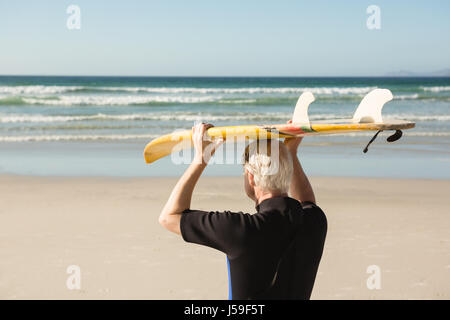 Vista posteriore del senior man tenendo la tavola da surf sulla testa in spiaggia durante il giorno di sole Foto Stock