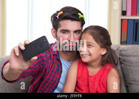 Padre indossa il make-up alla ricerca seria seduta con la figlia tenendo selfie Foto Stock