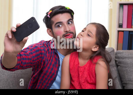 Padre indossa il make-up tirando facce buffe seduto con la figlia tenendo selfie Foto Stock