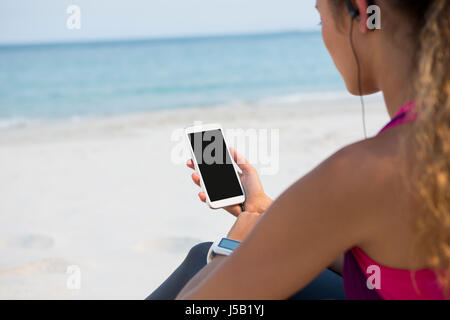 La sezione centrale della donna utilizzando il telefono cellulare mentre è seduto alla spiaggia contro sky Foto Stock