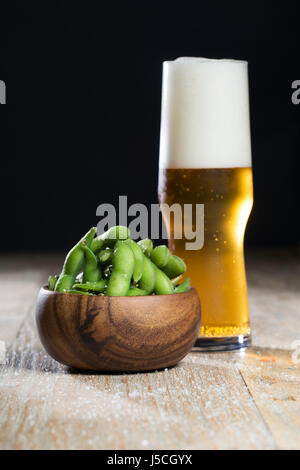 Ciotola di edamame e un bicchiere di birra seduti su una tavola in legno rustico. Foto Stock