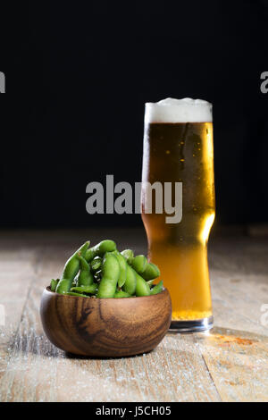 Ciotola di edamame e un bicchiere di birra seduti su una tavola in legno rustico. Foto Stock