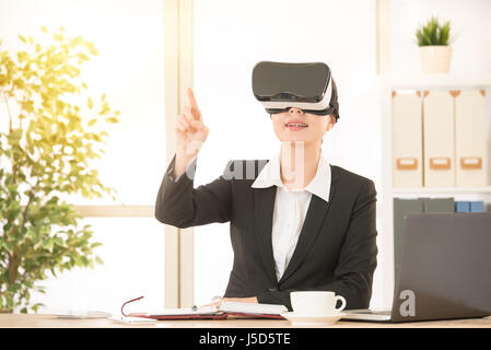 Eccitato giovane donna utilizzando un auricolare VR e puntamento touch screen su aria organizzare il contenuto del lavoro vivendo la realtà virtuale di lavoro video per l'ufficio. Foto Stock