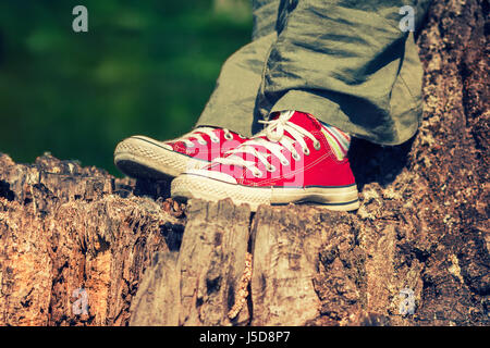 I piedi femminili in rosso brillante sneakers in tela, in piedi su un ceppo di albero, indossando verde pantaloni di lino Foto Stock