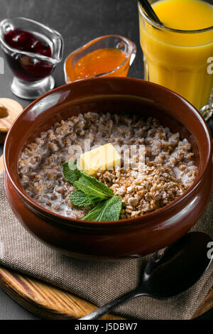 Farinata di grano saraceno con il latte in una ciotola su una tavola di legno. Prima colazione da farinata di grano saraceno, decorate con la menta e il burro. In background, succhi di frutta Foto Stock