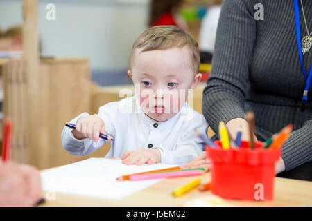 La sindrome di Down bambino facendo la colorazione in al suo vivaio. Il suo insegnante è seduta accanto a lui. Foto Stock