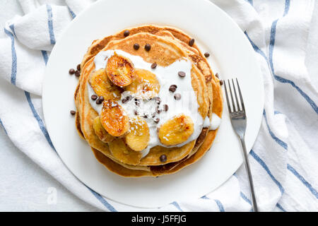 Pancake caramellato con banane, yogurt naturale, scaglie di cioccolato e scaglie di cocco sulla piastra bianca. Vista superiore Foto Stock