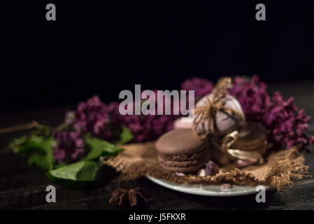 Caffè cremoso e macarons al buio su un tavolo di legno con lillà sullo sfondo Foto Stock