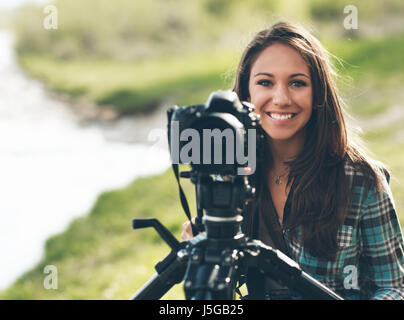 Donna sorridente fotografo utilizzando una telecamera digitale professionale su un treppiede, paesaggio naturale sullo sfondo Foto Stock