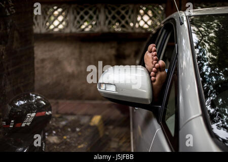 Un uomo addormentato nella sua auto con i suoi piedi fuori in Mumbai, India. Foto Stock