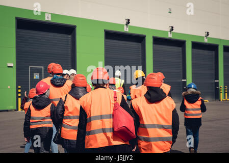 Un gruppo di persone in arancione caschi e giubbotti riflettenti visitando la fabbrica o il magazzino. Da dietro vista. Foto Stock