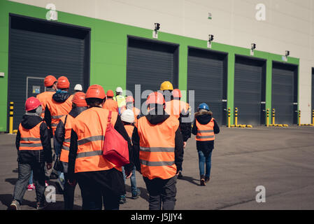 Un gruppo di persone in arancione caschi e giubbotti riflettenti visitando la fabbrica o il magazzino. Da dietro vista. Foto Stock