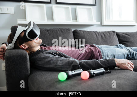Immagine del giovane uomo felice giace sul divano di casa indossando interni 3d occhiali per realtà virtuale. Foto Stock