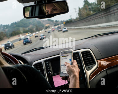 Driver femminile in l/h guidare il veicolo texting con iPhone smartphone durante la guida sulla trafficata autostrada californiana California USA Foto Stock