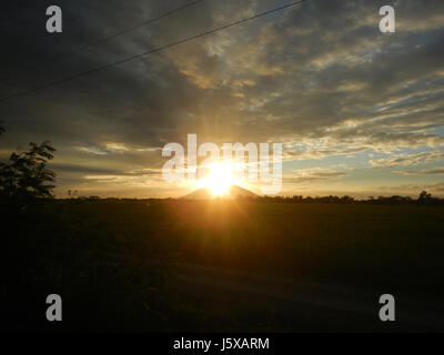 Questa immagine cattura la vista mozzafiato del Monte Arayat, situato a San Isidro, Nueva Ecija, Filippine. La fotografia presenta nuvole mistiche e gloriosi raggi di luce del tramonto, evidenziando la maestosità della montagna. Foto Stock