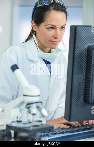 Un attenta femmina o medico ricercatore scientifico o medico donna utilizzando un computer in un laboratorio con microscopio e altre apparecchiature nel foreg Foto Stock