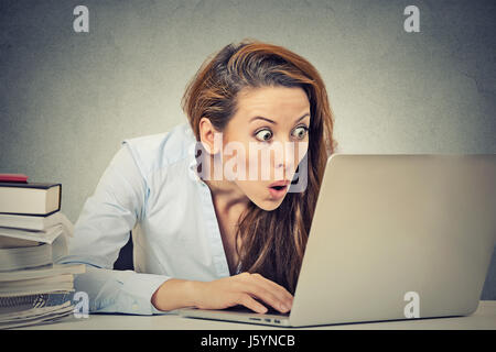 Ritratto di giovani scioccato business donna seduta nella parte anteriore del computer portatile che stanno guardando sullo schermo isolato muro grigio Sfondo. Funny Face emoti di espressione Foto Stock